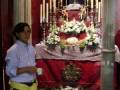 Una persona con una camisa azul y un cinturón amarillo, frente a un altar adornado con flores blancas y una imagen religiosa. La decoración es elaborada, con velas encendidas y un fondo rojo que contrasta con la blancaza de las flores.