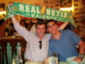 Dos hombres celebran con una bufanda del Real Betis Balompié, mostrando alegría y orgullo por el equipo.