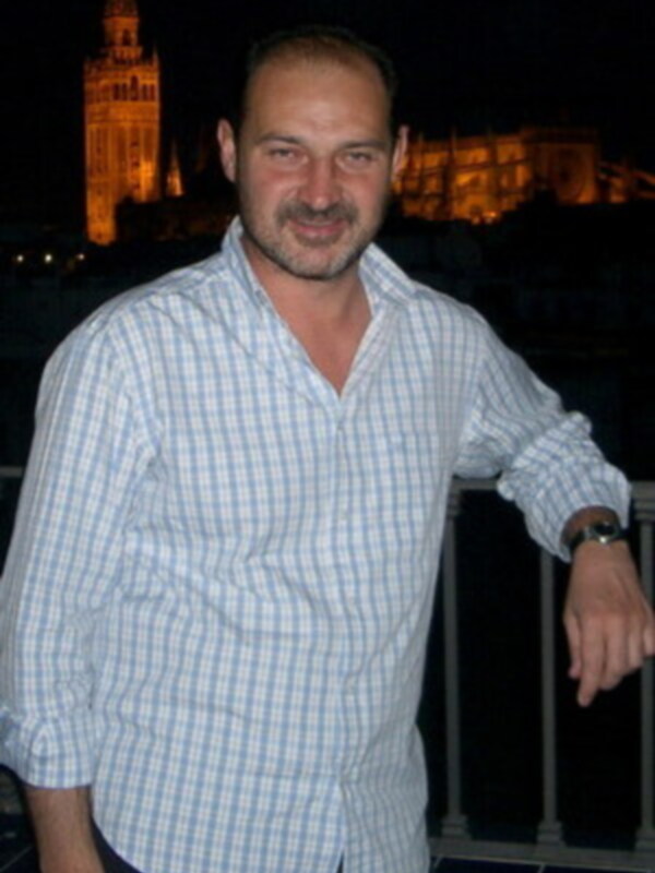 Un hombre con barba y bigote, vistiendo una camisa a cuadros azul claro y blanca, posa con el brazo en la barandilla de una terraza. En el fondo, se observa una torre iluminada en la noche y un edificio con luces de fondo.
