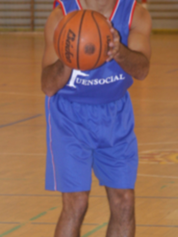 Un hombre en un uniforme de baloncesto azul y rojo, con la palabra "FuenSociales" en el pecho, sostiene un balón de baloncesto. Está en una cancha de baloncesto con líneas y marcadores visibles.