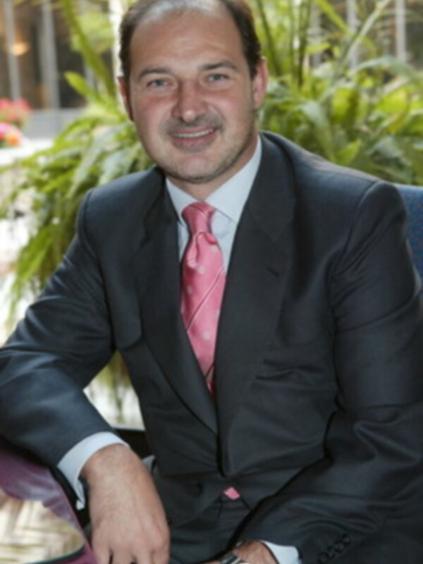 Un hombre con una camisa blanca y corbata rosa, posa en un entorno verde con plantas.