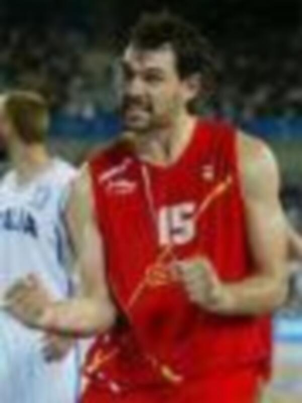 Un jugador de baloncesto en acción, con una camiseta roja y el número 15 visible. El fondo muestra a otros jugadores en un partido de baloncesto, con una cancha clara y sombras que indican luz solar.