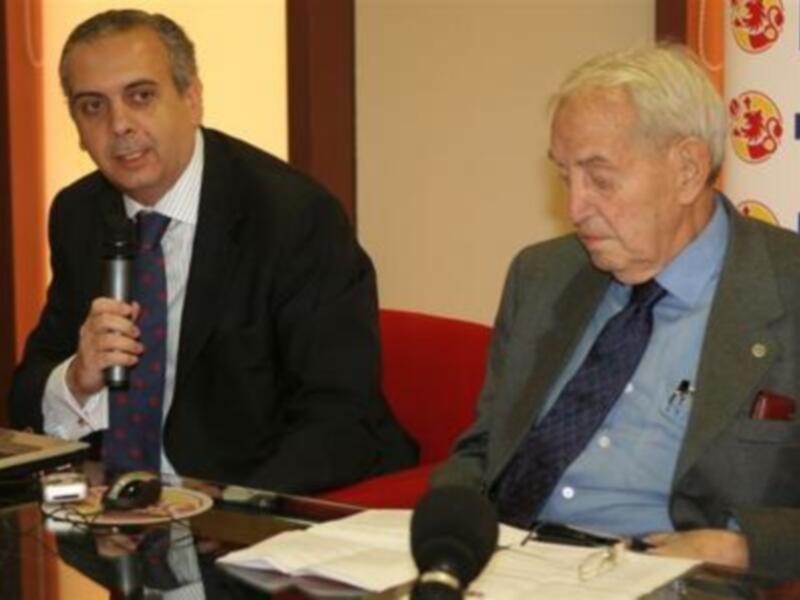 Dos hombres sentados en una mesa, uno con corbata y traje negro, el otro con corbata y chaqueta gris. La mesa tiene un plato de comida, una botella de agua y un micrófono. En el fondo, se ve una pared con carteles publicitarios.