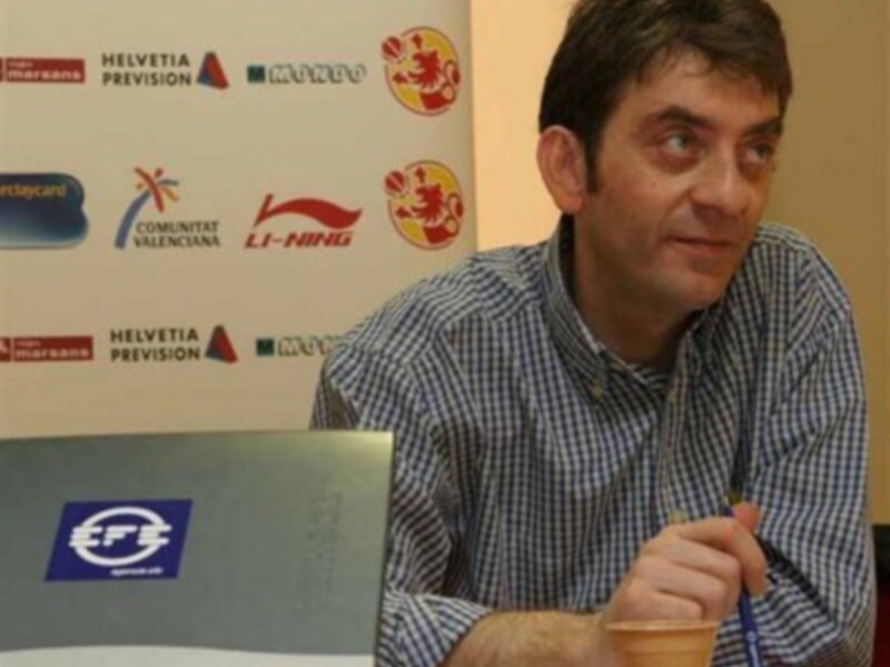 El hombre está sentado frente a una pared con varios logos de empresas, incluyendo Li-Ning y Helvetia. Está sosteniendo una taza de café con leche y un bolígrafo azul.