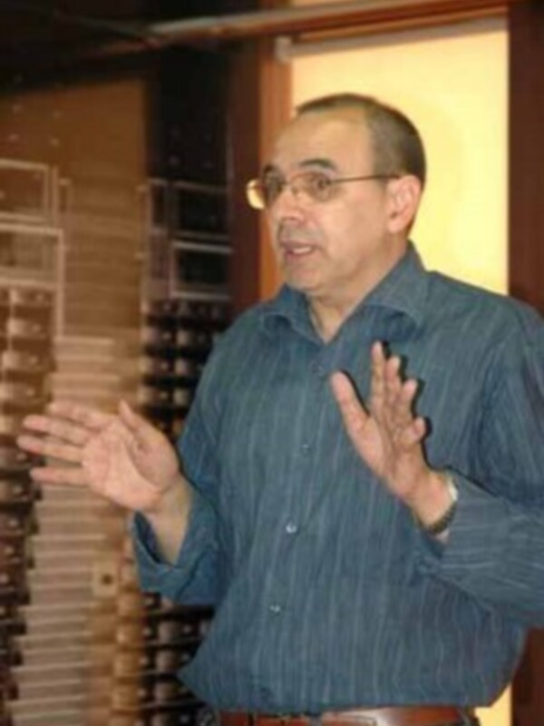 Un hombre en una camisa azul con rayas, hablando y gesturando con las manos. Parece estar en un lugar de trabajo o conferencia, con una pared de ventanas detrás.