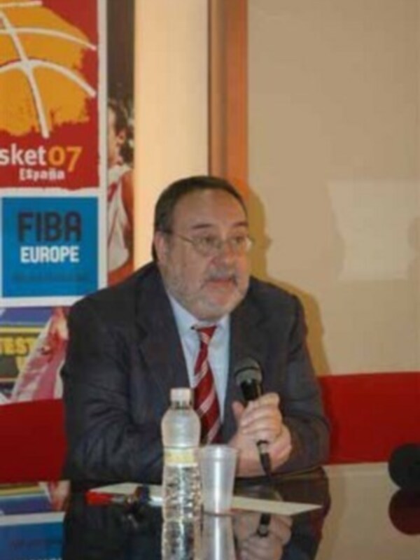 Un hombre en una conferencia de prensa, con una botella de agua y un micrófono en la mesa. En el fondo, hay una bandera de España y un cartel con la leyenda 'Basket 07'.