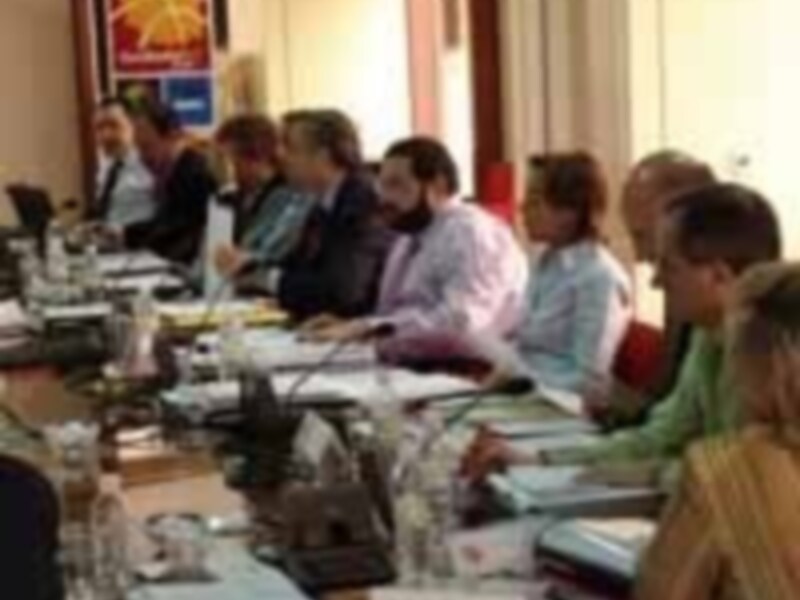 Una reunión en una sala de conferencias con varias personas sentadas frente a mesas llenas de documentos y computadoras. La pared está decorada con carteles y una bandera.