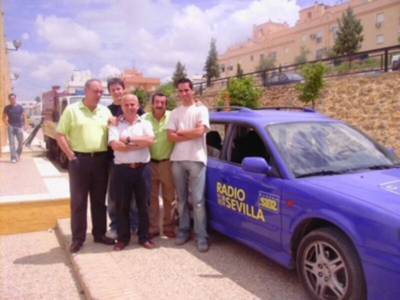 Un grupo de personas posando frente a un coche azul con la inscripción "Radio Sevilla". El entorno parece ser urbano, con edificios y árboles en el fondo.
