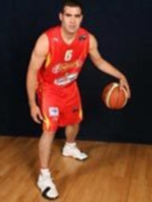 Un jugador de baloncesto con una camiseta roja y amarilla, pantalones cortos rojos y zapatillas blancas. El jugador sostiene un balón de baloncesto en su mano izquierda y está listo para jugar. El fondo es una cancha de baloncesto con líneas marcadas.