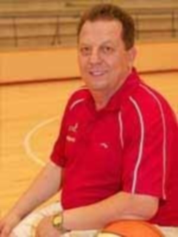 El entrenador de baloncesto está sentado en una cancha de baloncesto, con un jersey rojo y una camiseta deportiva. La imagen muestra su expresión seria, enfocada en la tarea del entrenamiento.