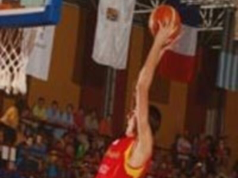 Un jugador de baloncesto realiza un tiro libre, con el aro y la canasta en primer plano. La escena se desarrolla en un estadio lleno de espectadores, con una bandera visible en el fondo.