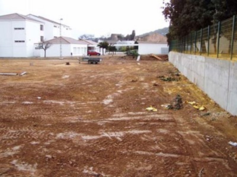 Un terreno baldío con vegetación seca y desgastada, rodeado por una valla de metal. En el fondo, edificios blancos y rojos se asoman entre árboles.