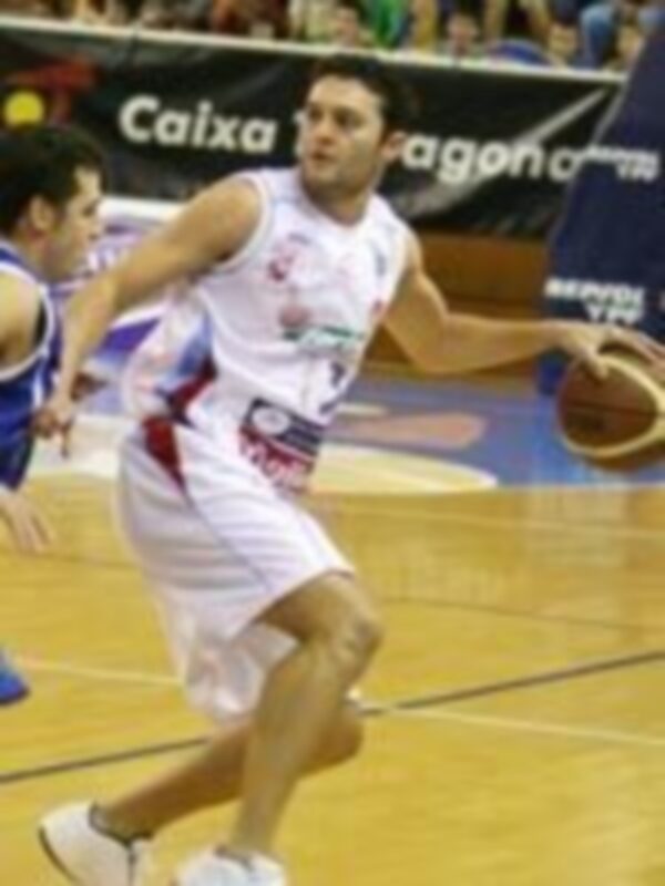 Un jugador de baloncesto en acción durante un partido, con el equipo local vestido en azul y blanco, y el equipo visitante en camiseta roja.