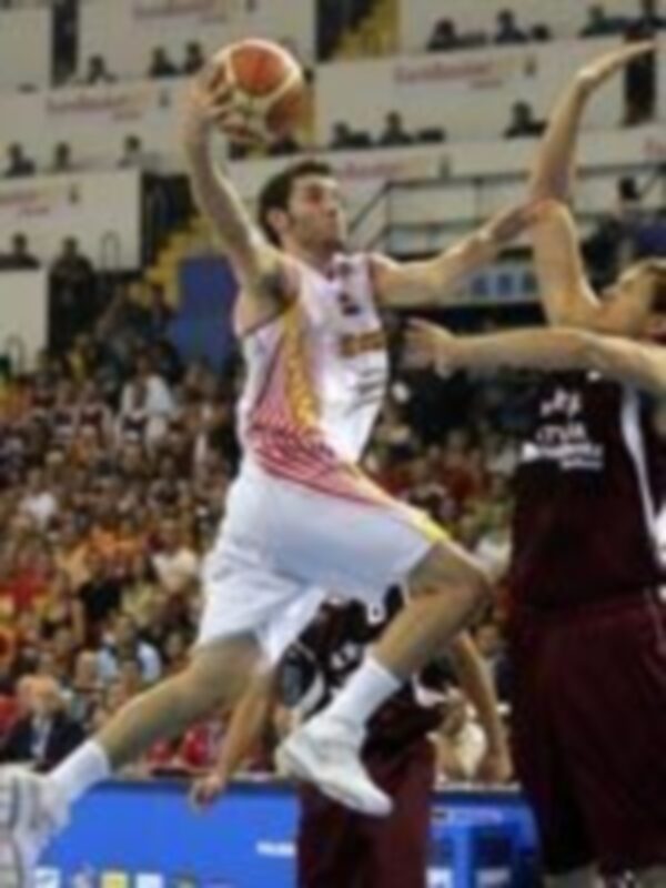 Un jugador de baloncesto en acción, lanzando el balón durante un partido. El fondo muestra a otros jugadores y la grada del estadio, con una iluminación brillante que resalta el momento de juego.