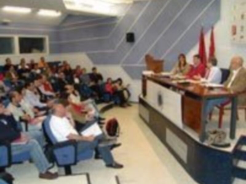 Una sala de conferencias con una gran pantalla en el fondo, personas sentadas en sillas alrededor de mesas y un hombre hablando frente a la pantalla.