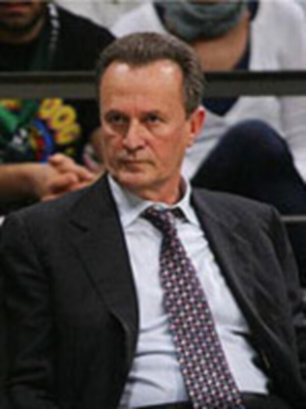 Un hombre con cabello oscuro y barba, lleva un traje negro y corbata roja. Está en una escena de deporte, posiblemente un partido de baloncesto, con otros jugadores en el fondo.