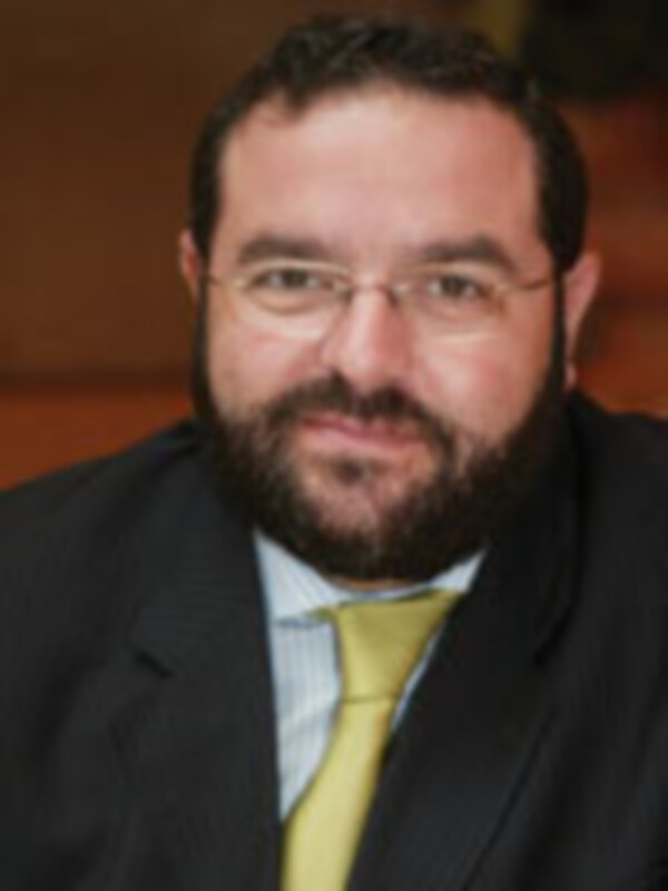 El hombre en la imagen lleva una camisa blanca con un cuello redondo, un chaleco amarillo y una corbata verde. Su cabello es oscuro y su barba es de color marrón.