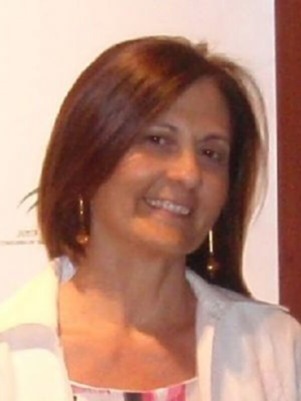 A una mujer con cabello corto y rojo, sonriendo. Se puede observar detalles como sus pendientes dorados y su vestimenta, que incluye una camisa blanca con detalles de colores.