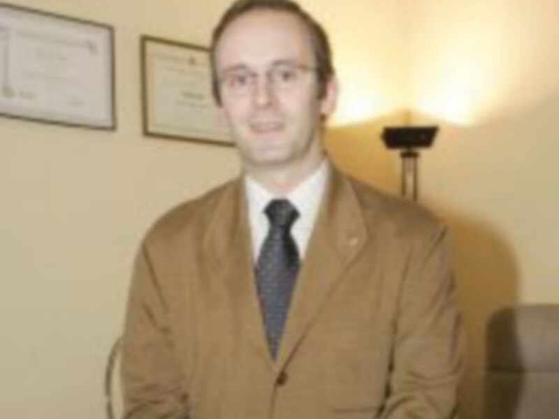 Un hombre con cabello corto y barba, vestido con una chaqueta beige y corbata azul, posa frente a un marco de fotos. La iluminación es suave y el fondo está oscuro, enfocando la atención en él.