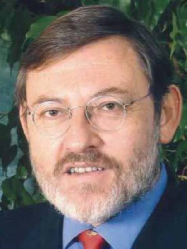 El hombre en la imagen lleva un traje oscuro, una camisa azul y una corbata roja. Está en un entorno natural con hojas verdes alrededor, lo que sugiere una conexión con la naturaleza.