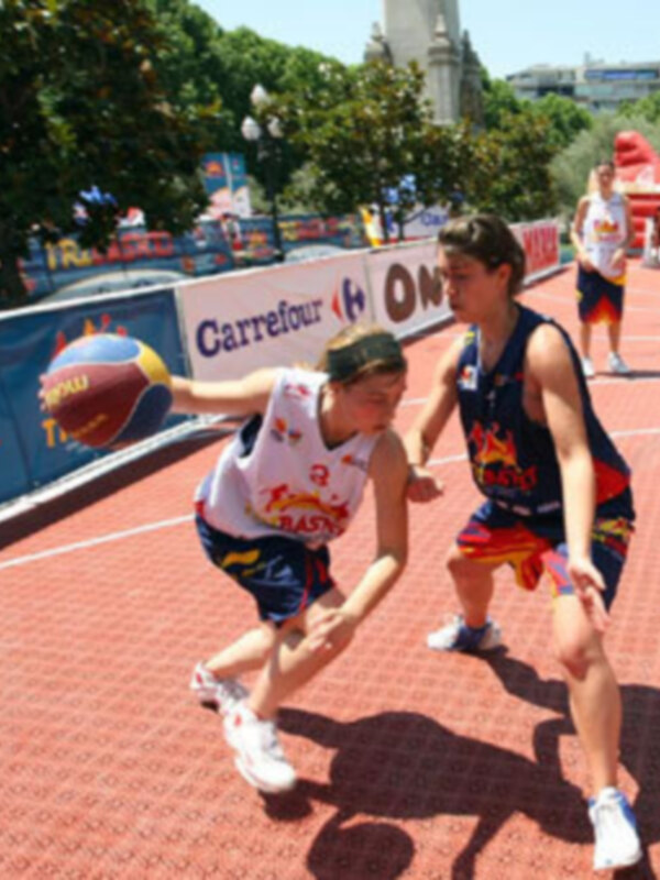 Dos atletas corren en una pista de atletismo, uno con un jersey blanco y otro con azul. La imagen está en un parque o estadio, con árboles y edificios al fondo.