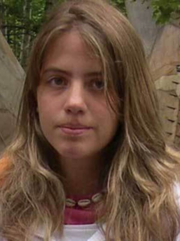 Una persona con cabello largo y ondulado, con una camiseta blanca y un collar de cuentas. Fondo con estructuras de madera y vegetación.