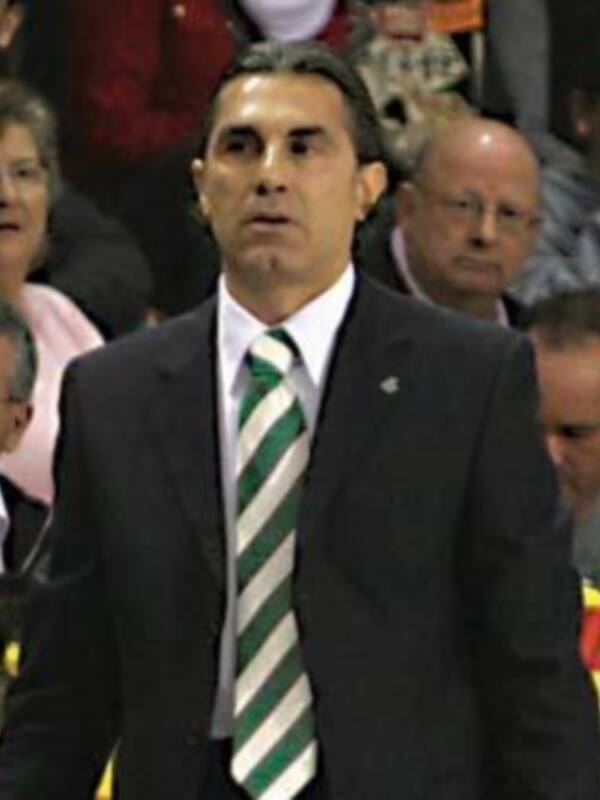 Un hombre en traje negro con corbata verde y una insignia en el pecho, mirando hacia la izquierda. En el fondo, hay personas con vestimentas informales y una pantalla de televisión en la parte superior derecha.