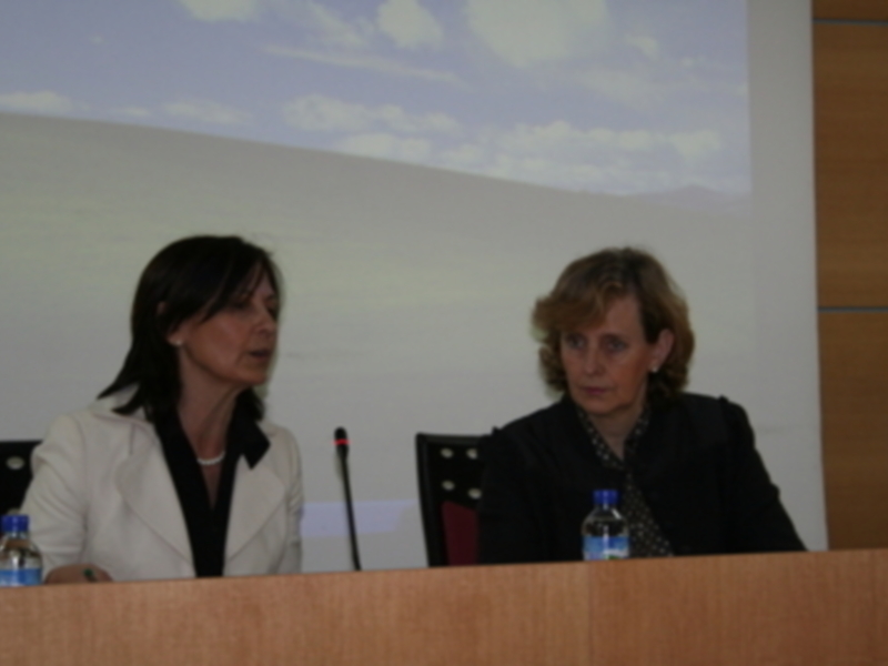 Dos mujeres participan en una conferencia en un escenario con una presentación de fondo de paisaje desértico.