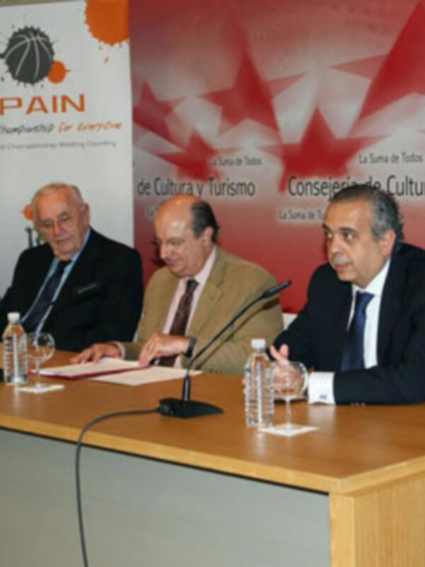 Tres hombres sentados en una mesa con micrófonos, frente a un cartel que dice "Spain". El texto relevante es "La Suma de Todos" y "Consejo de Cultura y Turismo".