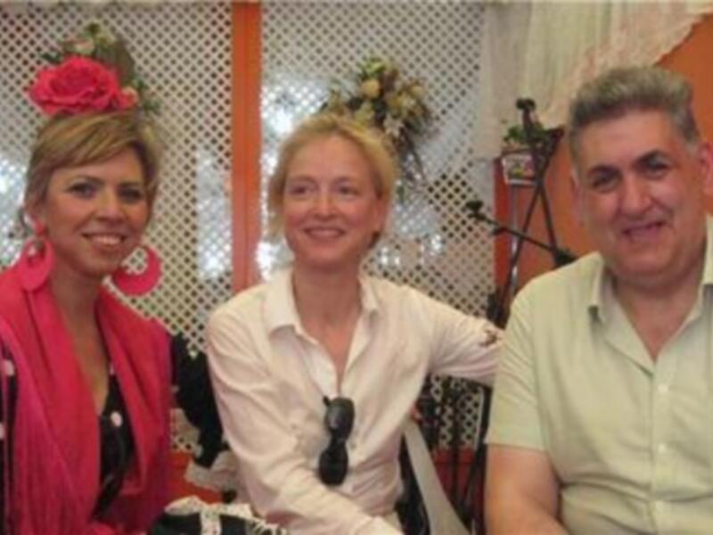 Una mujer con un vestido rojo y rosa, una flor en su cabello, sonríe junto a dos hombres. Detrás de ellos hay una ventana con un marco rojo y flores en el suelo.