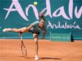 Una jugadora de tenis ejecuta una estocada hacia la pelota en un campo de tenis con el fondo que dice "Andalucía Marbella Club".