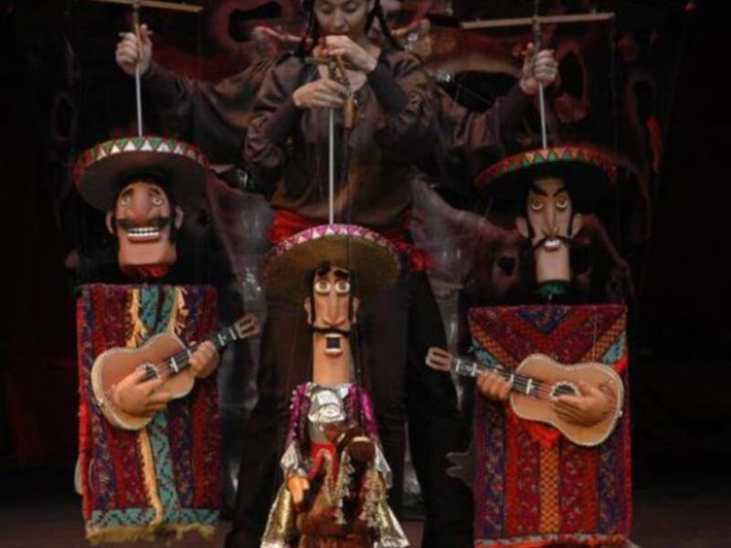 Una escena de marionetas mexicanas con personajes vestidos con sombreros de charro y mantas tradicionales, tocando instrumentos musicales.