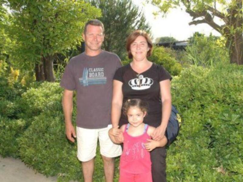 Una familia feliz en un parque, con un padre y una madre sonriendo para la cámara, mientras su hija les sostiene el brazo.