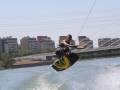 Un hombre realiza un salto de wakeboard en el agua, con una vista de edificios y un puente en el fondo.