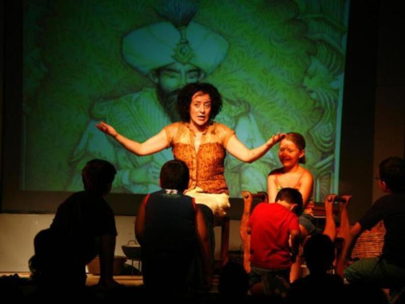 Una actriz en un vestido rojo se encuentra en el centro de la escena, con un fondo proyectado que muestra una imagen de un personaje antiguo. La escena parece ser parte de una representación teatral o una actuación.