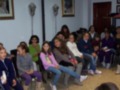 Grupo de niñas sentadas en sillas en una sala, posiblemente escuchando o participando en una actividad educativa o cultural.