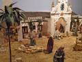 Escena navideña con figuras de personas y animales en un entorno rural. Hay una iglesia blanca con detalles dorados, un toldo y una casa en el fondo. La escena está ambientada en Sevilla.