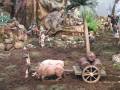 Escena navideña con figuras de madera en un paisaje montañoso. Un animal pequeño trilla la tierra mientras otro arrastra un carro con barriles de vino. En el fondo, figuras humanas y animales en una escena de natividad.