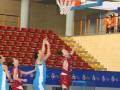 Jugadores de baloncesto en acción durante un partido en una cancha de baloncesto con asientos vacíos en el fondo.