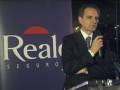 Un hombre en traje negro hablando al micrófono frente a un fondo azul con el logo de Realco Seguros.