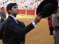 Un hombre en traje negro sostiene un sombrero de copa, posiblemente un matador de toros en una plaza de toros llena. El suelo es de arena y el público está sentado en gradas.