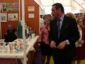 Un hombre en traje negro y corbata azul está presentando productos en una feria, con una mujer de fondo observando. En el fondo, se pueden ver carteles informativos y estantes con diferentes productos.