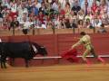 Un torero en traje de luces realiza una pasada con la capa roja frente a un toro en una plaza de toros, rodeado por un público entusiasmado.