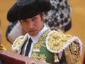 Un torero en traje tradicional con un sombrero negro y una chaqueta verde ornada de oro, sujetando la barandilla de un corral.
