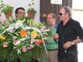 Tres personas, dos hombres y una mujer, posan frente a un gran ramo de flores. La imagen parece ser una foto de recuerdo o celebración, posiblemente en un evento formal. La decoración floral es impresionante y el ambiente sugiere una celebración o homenaje.