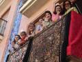 Grupo de mujeres en trajes tradicionales, posando desde un balcón con una tela bordada en el fondo.