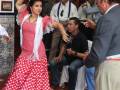 Una mujer en un vestido flamenco rojo y blanco baila al ritmo de la música mientras una persona toca el tambor. La escena se desarrolla en un ambiente festivo con decoraciones y carteles en el fondo.