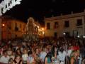 Una procesión nocturna en un pueblo con una gran imagen religiosa iluminada por faroles. La multitud, vestida de colores, observa atentamente la procesión mientras las banderas ondean en el aire. La escena es festiva y religiosa, con la luz de las farolas iluminando el ambiente nocturno.