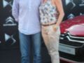 Un hombre y una mujer posan frente a un Citroën DS4 rojo en una presentación de automóviles. El hombre lleva un polo azul claro y pantalones vaqueros, mientras que la mujer usa una camiseta floral y pantalones beige. Ambos tienen las manos en los bolsillos de sus pantalones, y el hombre lleva una pulsera. En el fondo, se pueden ver los logos de Citroën y el DS4 rojo.