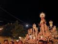 Una procesión religiosa nocturna con una imagen de la Virgen en el centro, rodeada por flores y luces. La luna se puede ver en el cielo nocturno.